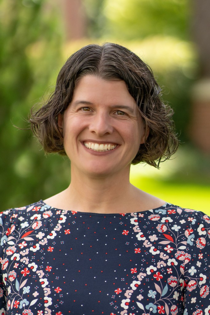 Headshot of a white woman with grayish-brown, curly, chin-length hair and brown eyes smiling.